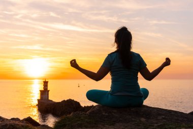 Gün batımında bir kayanın üzerinde meditasyon yapan bir kadın. Deniz fenerinin yanında, sağlıklı ve natürist bir yaşam sürüyor.