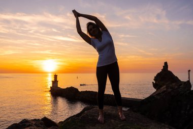 Sarışın bir kadın, gün batımında bir kayanın üzerinde deniz fenerinin yanında, sağlıklı ve natürist bir hayat sürüyor.