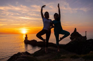 Vrksasana, gün batımında bir kayanın üzerinde meditasyon ve yoga yapan iki kadın, denizdeki bir deniz fenerinin yanında, sağlıklı ve natürist bir yaşam sürüyor.