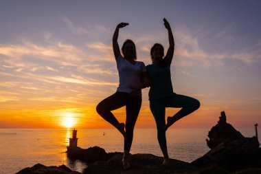 Vrksasana, gün batımında bir kayanın üzerinde meditasyon ve yoga yapan iki kadın, denizdeki bir deniz fenerinin yanında, sağlıklı ve natürist bir yaşam sürüyor.