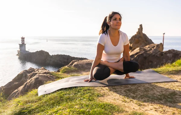 A Latin woman doing yoga exercises in nature by the sea, stretching next to a lighthouse