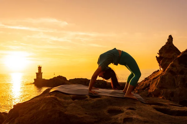 Urdhva Dhanurasana, gün batımında bir kayanın üzerinde meditasyon ve yoga egzersizleri yapan bir kadın. Denizde bir deniz fenerinin yanında, sağlıklı ve natürist bir yaşam sürüyor.