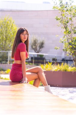 Alışveriş merkezinin önünde kırmızı elbiseli beyaz bir kız oturuyor.