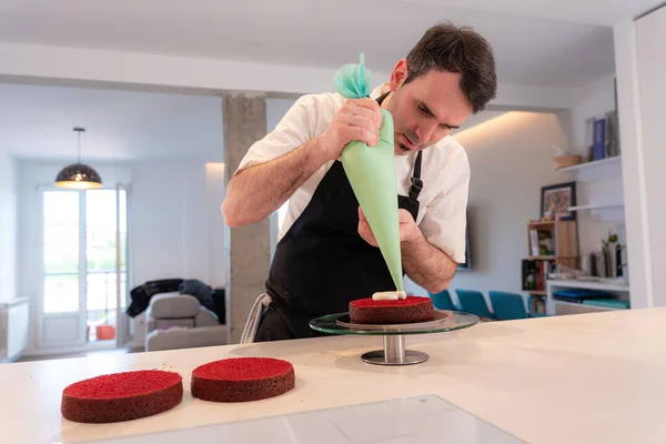 A challenger man cooking a red velvet cake at home, assembling the triple sponge base of the cake with the piping bag