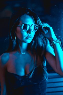 Geceleri şehirde mavi kollu, mavi gözlüklü ve saati olan esmer bir kız kameraya bakıyor.