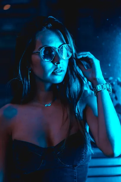 Brunette model at night in the city with blue leds, blue glasses and a watch, looking at the camera