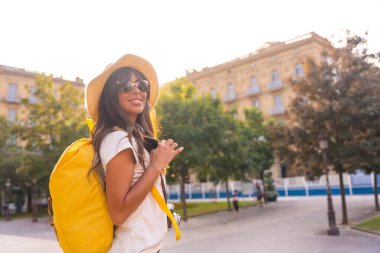 Yaz günbatımında şapkalı bir turist. Fotoğraf makinesi, şapka, sarı sırt çantası ve güneş gözlüğü olan bir kilisenin yanında.