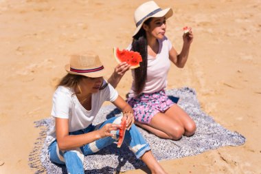 Yazın kumsalda arka planda deniz olan bir karpuz yiyen, kumların üzerinde oturan iki arkadaş.