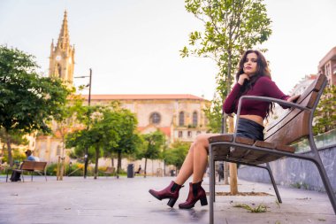 Deri etekli esmer bir kadın sonbaharda arka planda bir kiliseyle şehirde dinleniyor.