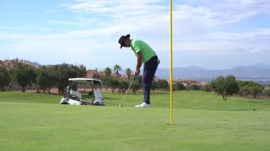 Golf oynayan bir adam, golf sopasıyla topu yeşildeki deliğe atıyor.
