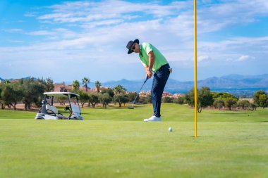 Golf oynayan bir adam, golf sopasıyla topu yeşildeki deliğe atıyor.