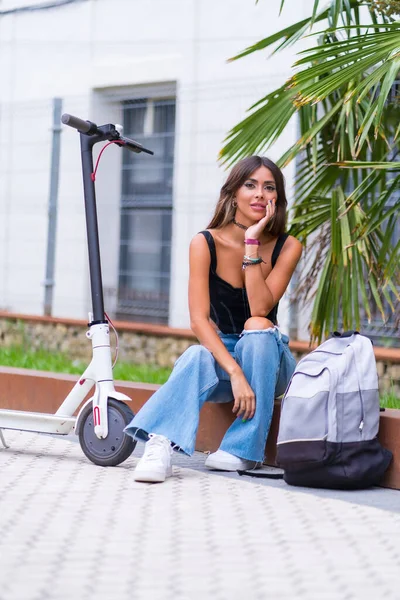 Portrait of a young woman sitting in the city waiting for friends with an electric scooter