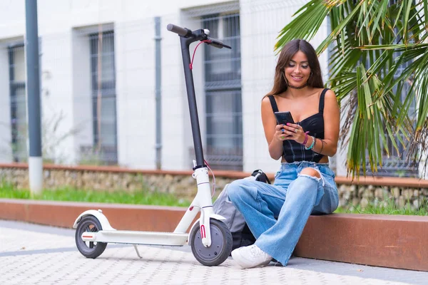 Portrait of a young woman sitting in the city with an electric scooter and looking at social networks