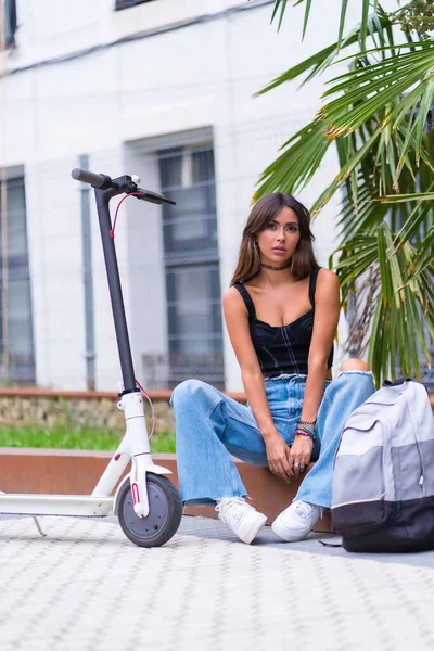 Portrait of a young woman sitting in the city waiting for friends with an electric scooter