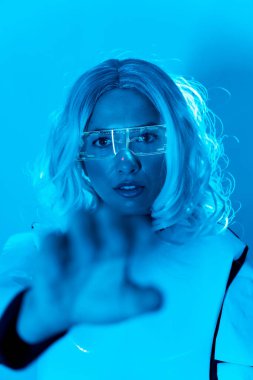 Fütüristik kavram, mavi arka planda aydınlanmış sanal gerçeklik gözlükleriyle elini uzatan genç bir kadın.