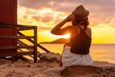 İbiza adasındaki Cala Comte plajında gün batımında şapkalı genç bir kadın. Balearic