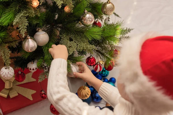 Child boy in red Santa hat decorates Christmas tree close up. Xmas or New Year cozy holidays concept. Selective focus — Stock Photo, Image