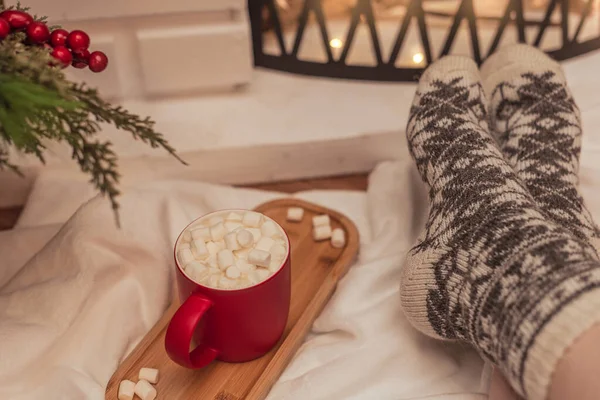 Sıcak titreşimler sahnesi, beyaz marşmelovlu bir fincan sıcak içecek ve şöminenin önünde sıcak yün çoraplı kadın ayakları. Seçici odaklanma. Kış tatili konsepti boyunca rahat atmosfer — Stok fotoğraf