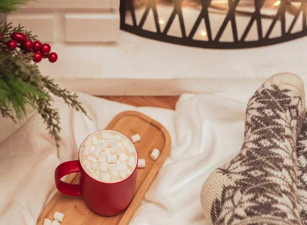 Cozy vibes scene with a cup of hot drink with white marshmallows and female feet in warm woollen socks in front of fireplace. Selective focus. Cozy atmosphere during winter holidays concept — Stock Photo, Image