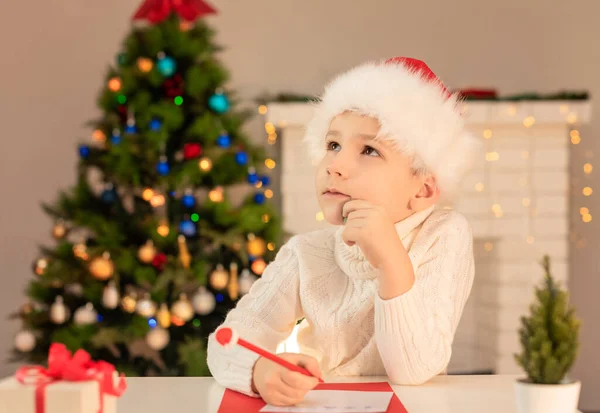 Child boy in red Santa hat writing a letter to Santa Claus. Christmas or New Year cozy holidays concept. Xmas time. Selective focus — Stock Photo, Image