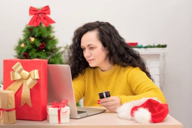 Arka planda Noel ağacına karşı bilgisayarında kredi kartı ve alışveriş yapan mutlu bir kadın var. Xmas çevrimiçi alışveriş, yeni yıla hazırlık, hediye alma, kış tatili satış konsepti