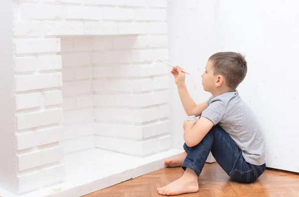 Niño se sienta en el suelo y pinta una chimenea decorativa con gouache de pintura blanca. Mejora del hogar, concepto de bricolaje. Enfoque selectivo Imágenes De Stock Sin Royalties Gratis