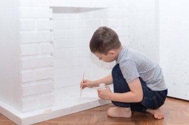 Çocuk yere oturur ve beyaz boya kebabıyla dekoratif bir şömine boyar. Ev geliştirme, DIY konsepti. Seçici odak
