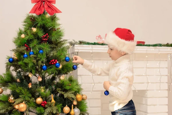 Chłopiec w czerwonym kapeluszu Mikołaja dekoruje choinkę z bliska. Boże Narodzenie lub Nowy Rok przytulny pomysł na wakacje. Skupienie selektywne — Zdjęcie stockowe