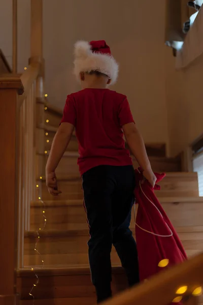 Little boy in Santa hat carries a bag of gifts up the stairs. Christmas or Happy New Year concept. Xmas spirit — Stock Photo, Image