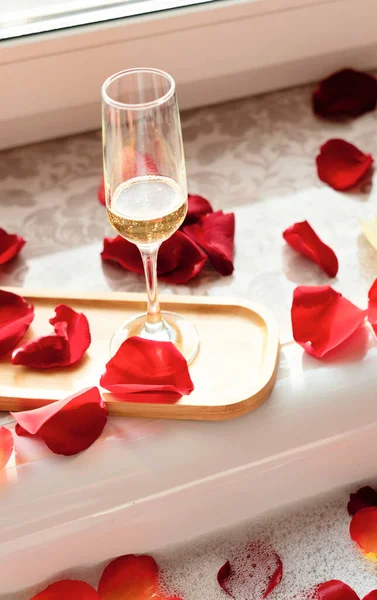 Glass of white wine near a bathtub with foam and red rose petals against a window. Enjoying life concept — 스톡 사진