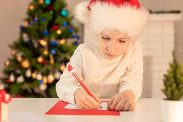 Child boy in red Santa hat writing a letter to Santa Claus. Christmas or New Year cozy holidays concept. Xmas time. Selective focus — Stock Photo, Image