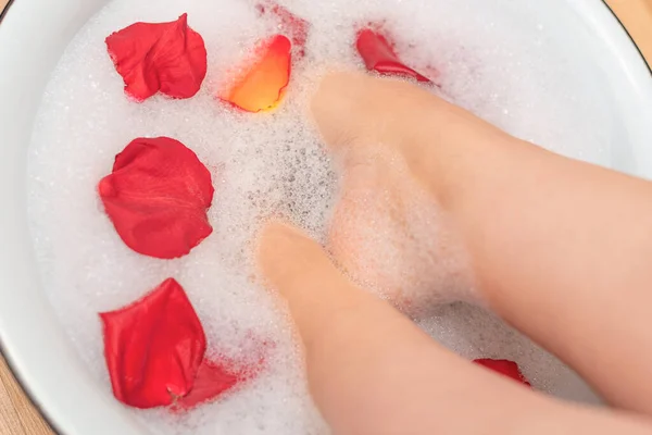 Woman feet in water with soap foam and rose petals close up with selective focus. Spa treatment for dry feet skin. Homemade bath soak for female feet and hand. Skin care concept. Luxurious lifestyle
