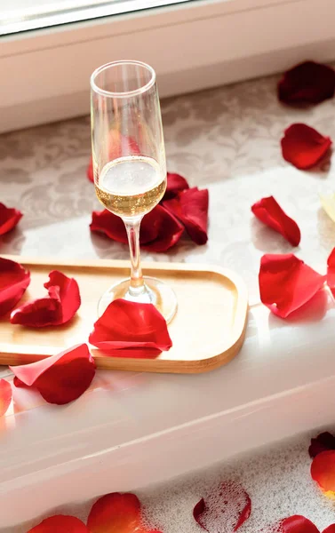 Glass of white wine near a bathtub with foam and red rose petals against a window. Enjoying life concept 스톡 사진