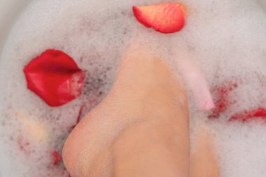 Sabun köpüğü ve gül yapraklı kadın ayakları seçici bir odaklanma ile yaklaşır. Kuru ayak derisi için spa tedavisi. Kadın ayakları ve elleri için ev yapımı banyo. Deri bakımı konsepti. Lüks yaşam tarzı.