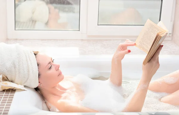 Young woman takes a bath with lush foam near the window reading the book. Personal hygiene, healthcare, relax at home concept — Φωτογραφία Αρχείου