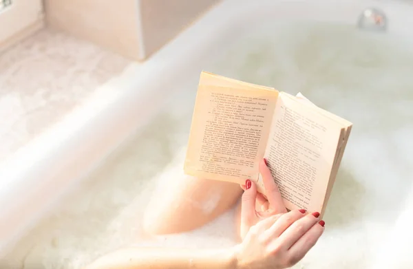 Young woman takes a bath with lush foam near the window reading the book close up. Personal hygiene, healthcare, relax at home concept. Selective focus — Φωτογραφία Αρχείου