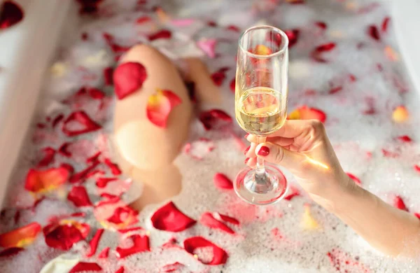 泡と赤いバラの花びらと浴槽に座っている間、女性の手は白ワインのガラスを保持します。人生の概念を楽しむ。選択的焦点 — ストック写真