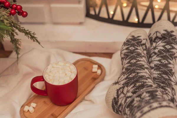 Sıcak titreşimler sahnesi, beyaz marşmelovlu bir fincan sıcak içecek ve şöminenin önünde sıcak yün çoraplı kadın ayakları. Seçici odaklanma. Kış tatili konsepti boyunca rahat atmosfer — Stok fotoğraf