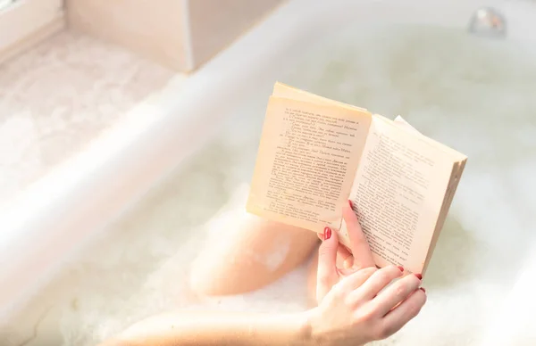 Young woman takes a bath with lush foam near the window reading the book close up. Personal hygiene, healthcare, relax at home concept. Selective focus — Φωτογραφία Αρχείου