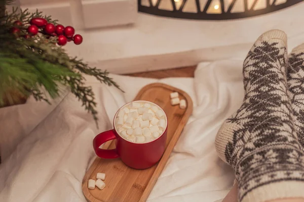 Ambiente acogedor con una taza de bebida caliente con malvaviscos blancos y pies femeninos en calcetines de lana caliente frente a la chimenea. Enfoque selectivo. Ambiente acogedor durante las vacaciones de invierno concepto —  Fotos de Stock