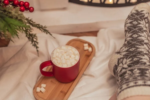 Ambiente acogedor con una taza de bebida caliente con malvaviscos blancos y pies femeninos en calcetines de lana caliente frente a la chimenea. Enfoque selectivo. Ambiente acogedor durante las vacaciones de invierno concepto —  Fotos de Stock