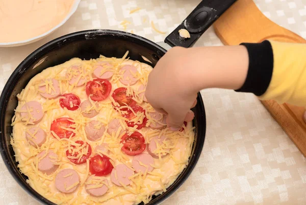 Cârnați, roșii și brânză plasate pe aluatul crud pentru pizza de casă de mână de copil în tigaie neagră. Vedere de top cu focus selectiv. Procesul de gătit pizza, pasul 9 — Fotografie, imagine de stoc