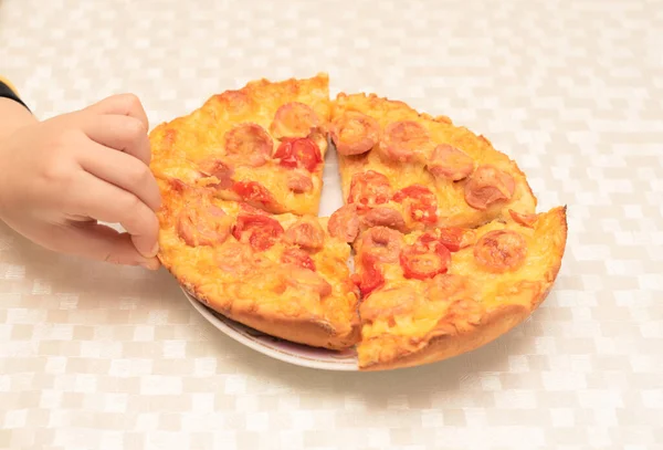 Childs mão leva um pedaço de pizza caseira com salsichas, tomates e queijo na mesa na cozinha de perto com foco seletivo. Processo de cozinhar pizza, passo 14 — Fotografia de Stock
