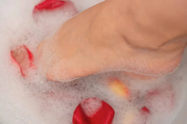 Woman feet in water with soap foam and rose petals close up with selective focus. Spa treatment for dry feet skin. Homemade bath soak for female feet and hand. Skin care concept. Luxurious lifestyle