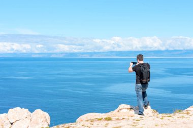 Sırt çantalı bir adam dünyanın sonundaki bir kayanın üzerinde duruyor ve gölün güzelliğini fotoğraflamak için akıllı telefonunu kullanıyor. Yürüyüş ya da seyahat konsepti. Açık havayı keşfediyorum. Doğanın güzelliği