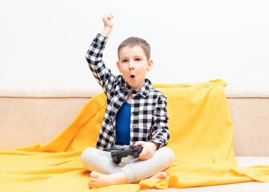 Çek gömlekli çocuk elinde siyah joystick ile kanepede oturmuş video oyunu oynuyor. Ev konseptinde video oyunları oynamak