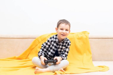 Çek gömlekli mutlu çocuk elinde siyah joystick ile kanepede oturmuş video oyunu oynuyor. Ev konseptinde video oyunları oynamak