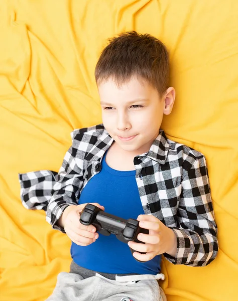 Menino feliz em camisa verificada deitado no sofá com joystick preto em suas mãos jogando o jogo de vídeo. Jogar jogos de vídeo em casa conceito — Fotografia de Stock