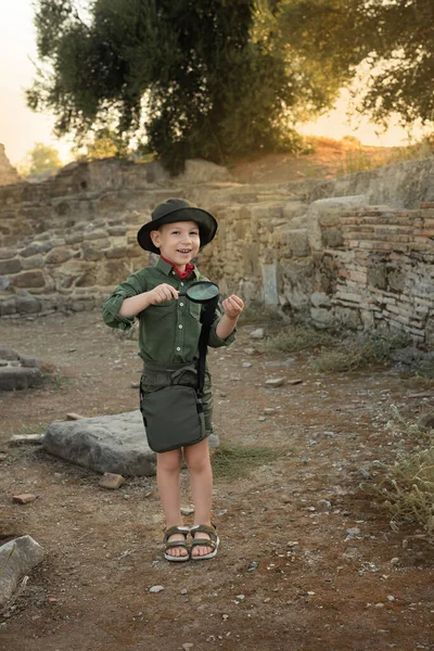 Menino arqueólogo em roupas cáqui estudando algo com lupa entre as ruínas de uma cidade antiga. Conceito de emprego diverso e não tradicional Imagens Royalty-Free