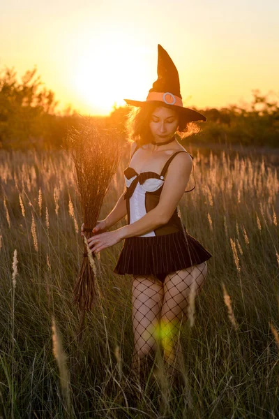 Sexy young woman in a witch\'s hat and a broom in a field, against the backdrop of a sunset. Concept for halloween.